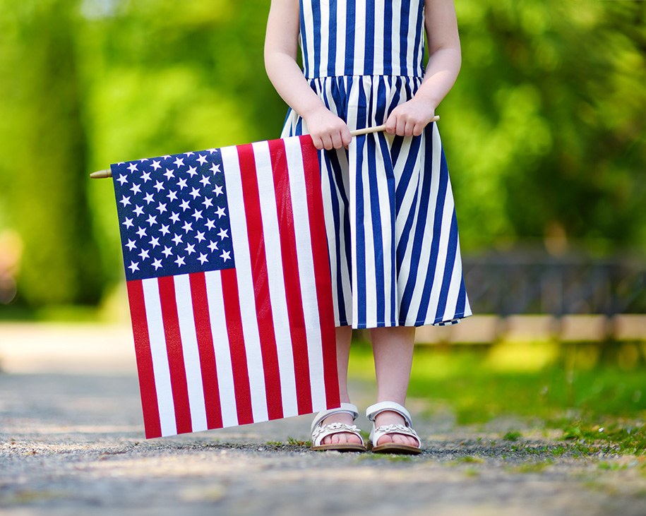 A little girl wearing a white and blue striped dress and white sandals holding a United States flag