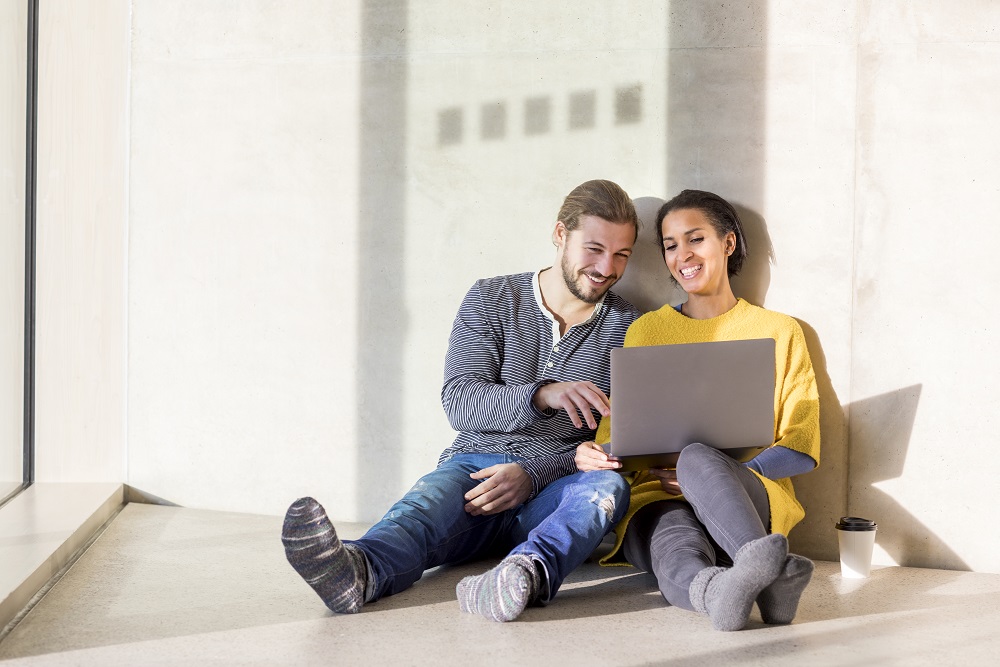 En la página, 2024. Pareja joven sentada en el suelo que ríe mientras usa una computadora portátil