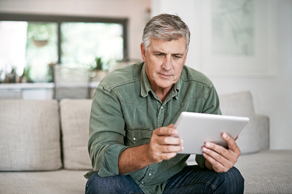 En la página, 2024. Un hombre de mediana edad sentado en un sofá mirando seriamente una tableta en sus manos.