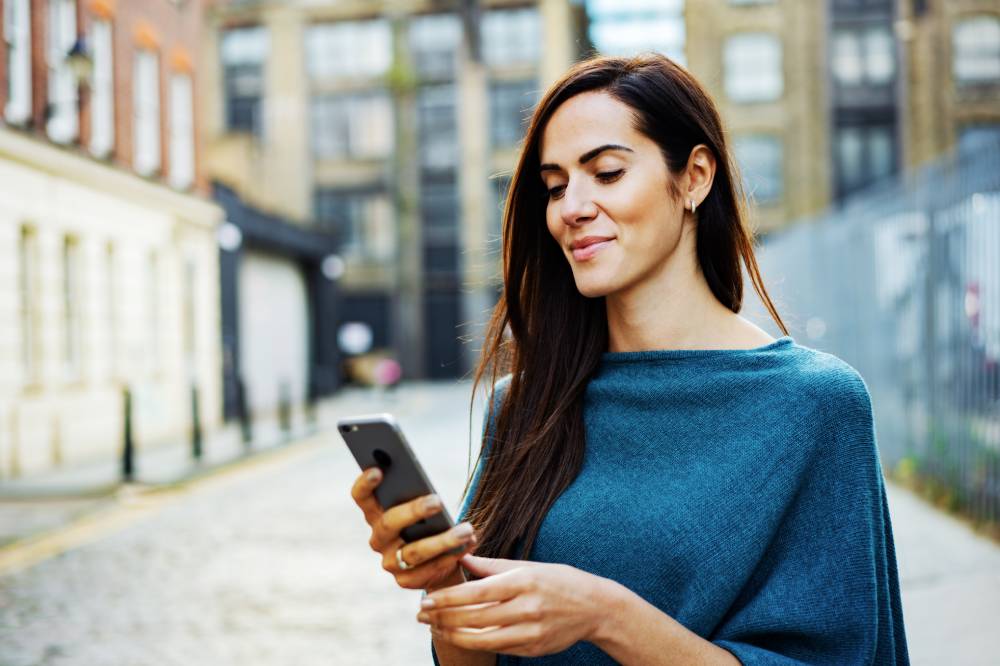 En la página, 2024. Una mujer parada al aire libre en una ciudad sostiene su teléfono y sonríe levemente a la pantalla.