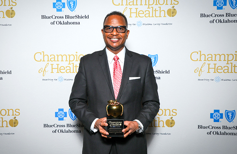 Man in a suit and tie holds a trophy in front of a backdrop with logos of Blue Cross and Blue Shield of Oklahoma and Champions of Health