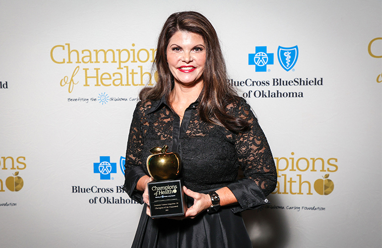 Woman in formal dress holds a trophy in front of a backdrop with logos of Blue Cross and Blue Shield of Oklahoma and Champions of Health