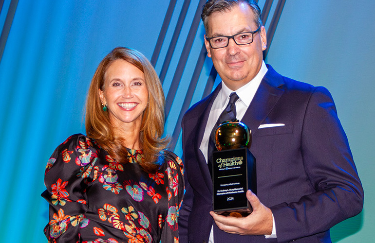 Man in suit and tie and glasses holds an award standing next to a women in floral print dress