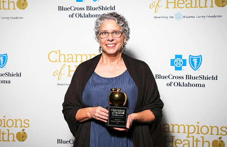 Woman in formal dress holds a trophy in front of backdrop with logos of Blue Cross and Blue Shield of Oklahoma and Champions of Health