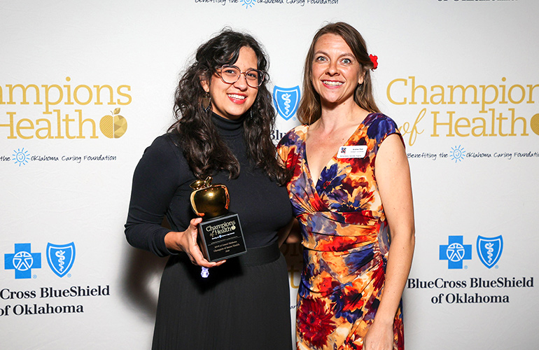 Two women in dresses hold a trophy in front of a backdrop with the logos of Blue Cross and Blue Shield of Oklahoma and Champions of Health